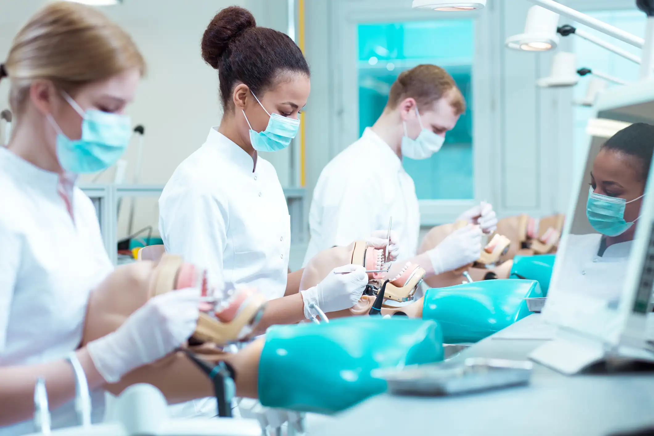 Four masked dental students in white coats practicing procedures on manikin heads at teal-topped training stations in a lab