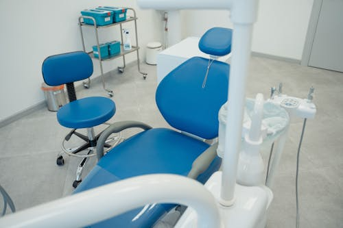 A dental treatment room with a blue patient chair and stool, a supply cart, and a dental delivery unit with handpieces